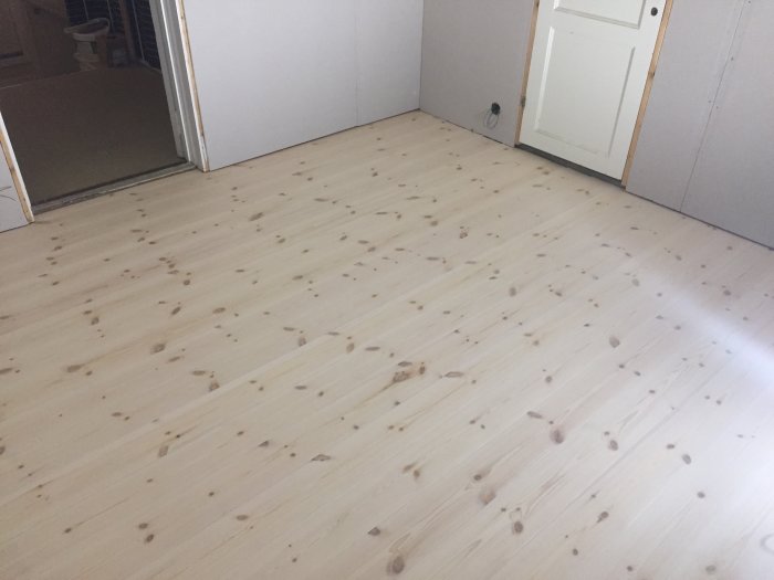 Nylagda obehandlade furugolvplank i ett ljust gästrum, med öppen dörr.