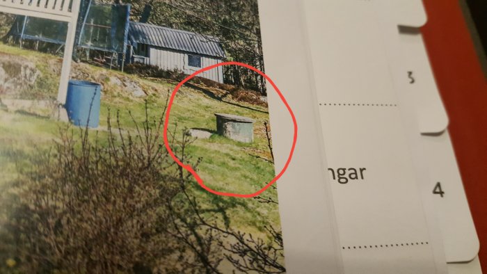 Brunn med lock placerad på gräsmark nära hus, markerad med röd cirkel.