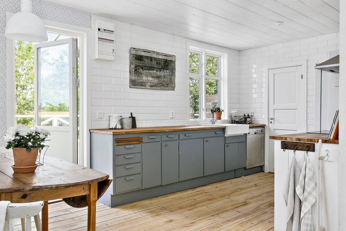 Ljus köksinteriör med vita väggar, grå skåp, träbänkskiva och -golv, samt ett träbord med blommor.