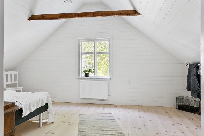 Ljus sovrumsmiljö med snedtak, vita väggar, träbjälke och trägolv, enkelt inredd med säng och fönster.