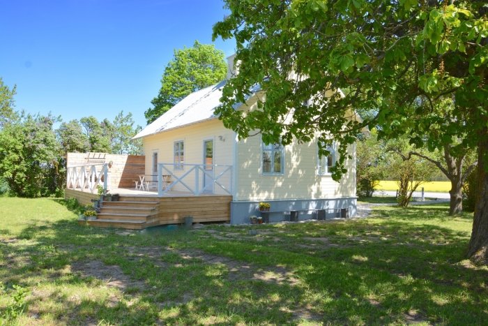 Ljust gult hus med vit veranda och trätrall i lummig trädgård under klarblå himmel.