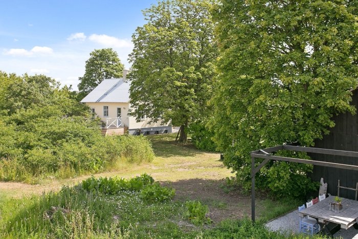 Lantlig vy med ett gult hus omgivet av gröna träd, en gräsmatta och en träterrass med bord och stolar.