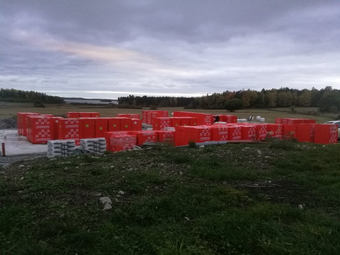 Staplar av röda levererade byggstenar på en lerig byggarbetsplats i skymningen.