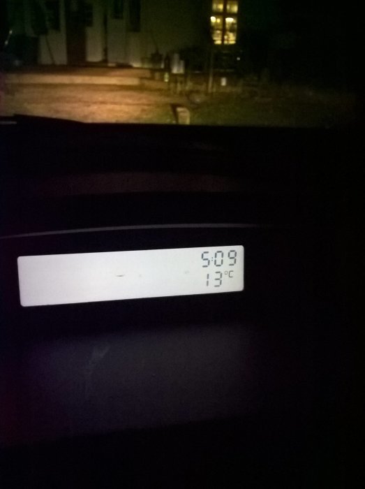 Bild av en termometer som visar 13 grader Celsius kl. 5:09 mot en suddig bakgrund av ett hus och trädgård på kvällen.