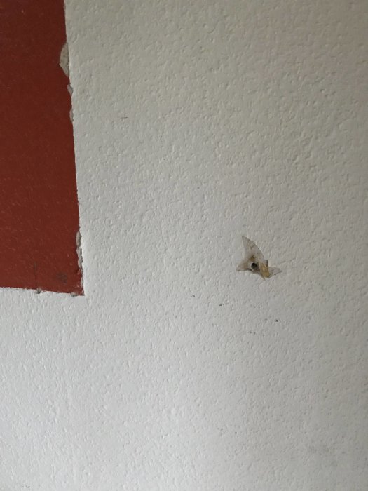 Vägg med strukturtapet och en delvis bortriven yta avslöjar underliggande rött lager.