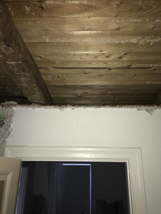 Synligt bjälklag ovanför ett fönster i ett rum under renovering, med ett gap mellan tak och vägg.