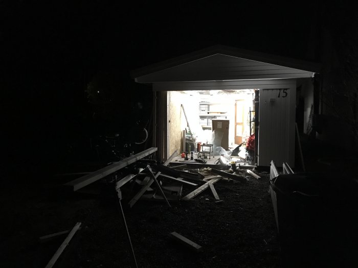 Garage med öppen port och material spridda på marken på kvällen, dåligt upplyst arbetsplats.