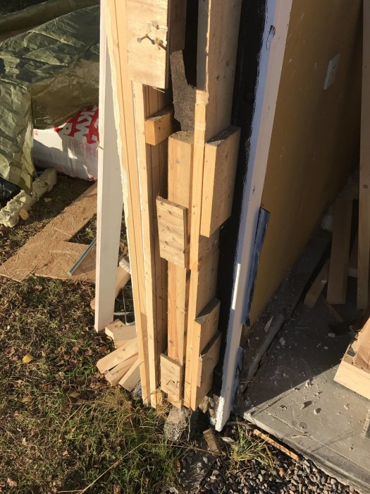 Oorganiserad hög av träplankor och byggmaterial vid en husvägg under renovering.