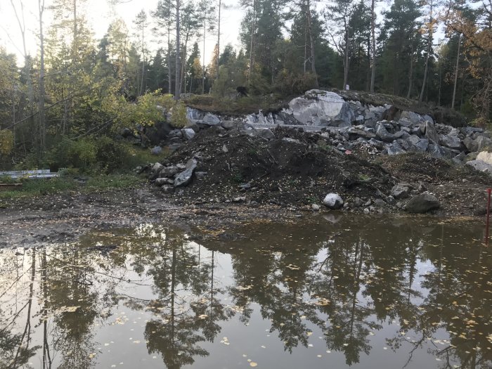 Vattenansamling vid husgrund med reflektion av träd, grushög i bakgrunden.