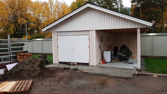 Garage med nymonterade vindskiveplåtar och en böjd plåt över nockpannan vid taket, jordhög och skottkärra i förgrunden.