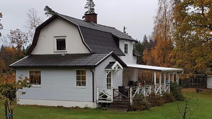 Tvåvåningshus med vit fasad och svart tak, delvis täckt av lövträd i höstfärger, med en veranda.