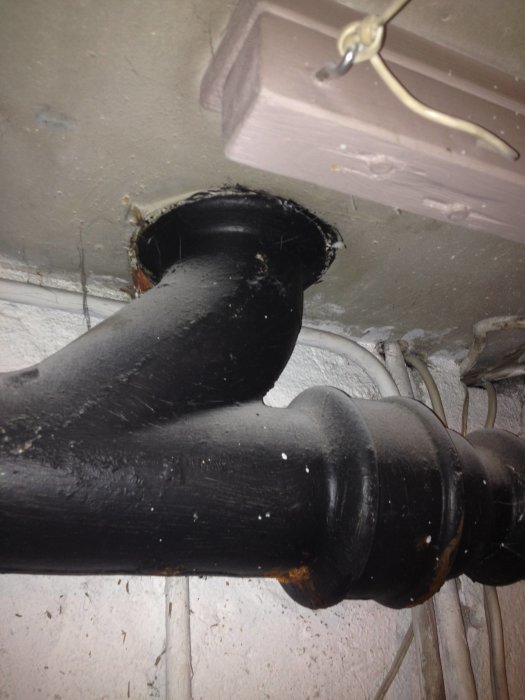 Rostigt gjutjärnsrör med en krök som går in i bjälklaget, synlig rost och gammal isolering runt genomföringen.