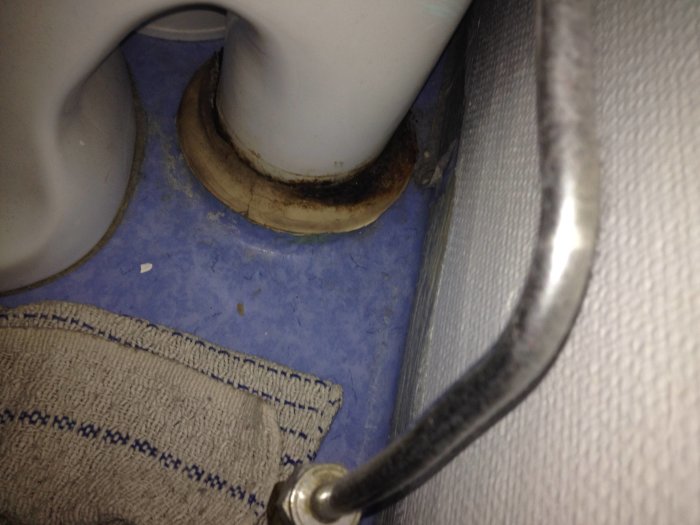 Rostig avloppsmuff vid toalettstosens fot på ett flytspacklat golv med blå plastmatta och detalj av infästning.