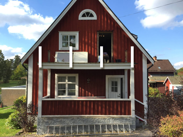 Röd stuga med vita stolpar och hängränna mellan, öppen veranda och grönska runt om.