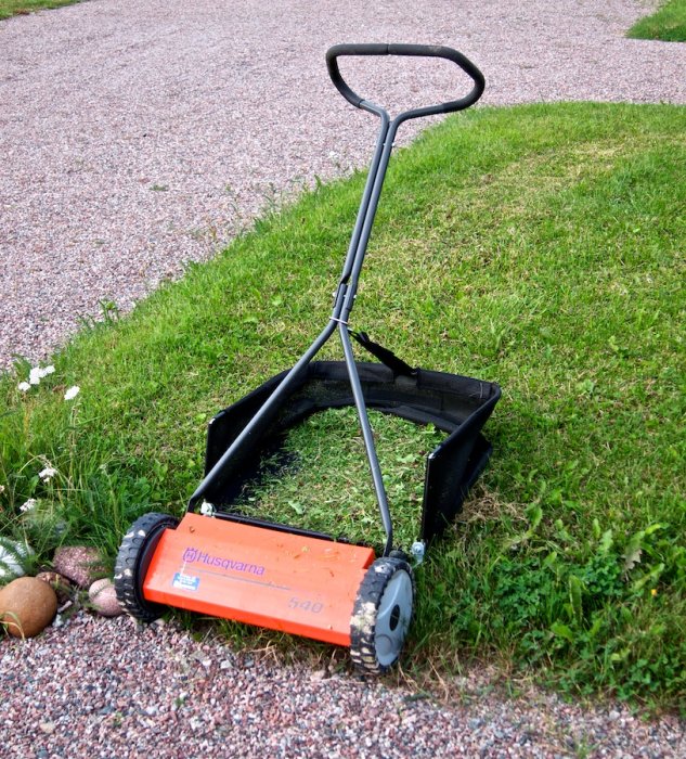 Manuell gräsklippare av märket Husqvarna på ett gräsmatta nära grusväg.