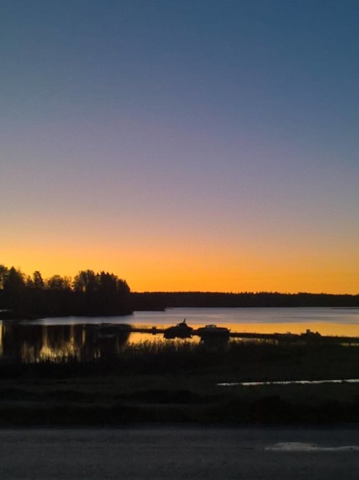 Soluppgång över en frusen sjö med silhuetter av träd och en brygga, himlen med nyanser av orange och blått.