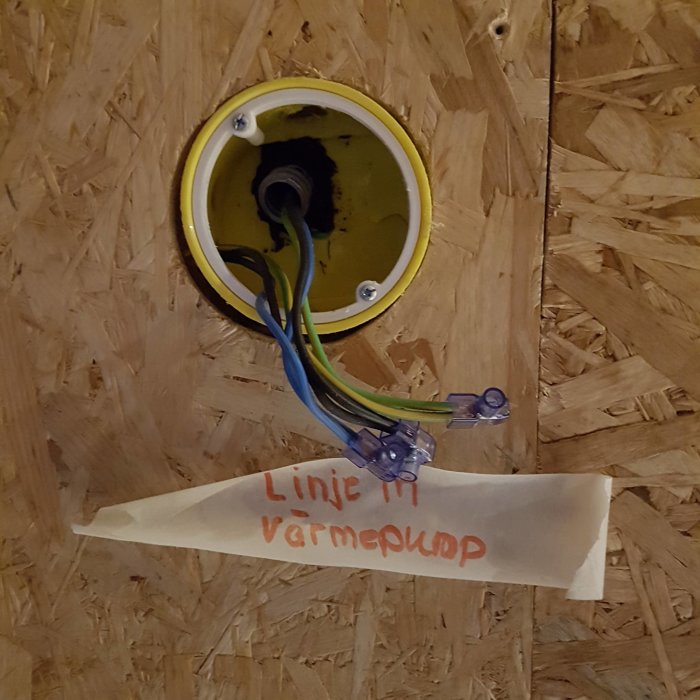 Öppen kopplingsdosa med blå, brun och gul/grön elkabel och etiketten "Linje till värmepump" på trävägg.