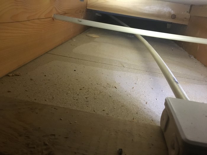 Dammigt utrymme under en säng med synliga elkablar och träpaneler.