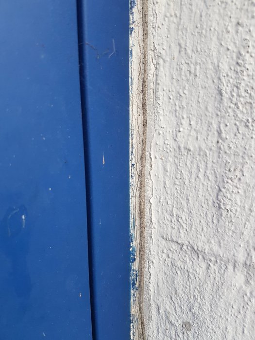 Sprucken mjukfog mellan blå dörrkarm och putsad tegelfasad.
