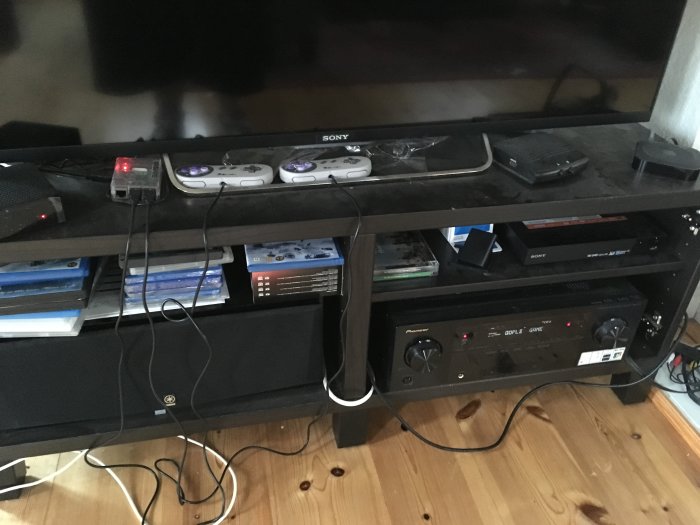 Hembioanläggning med receiver och spelkonsoler på en svart TV-bänk, synliga kablar och Blu-ray-filmer.