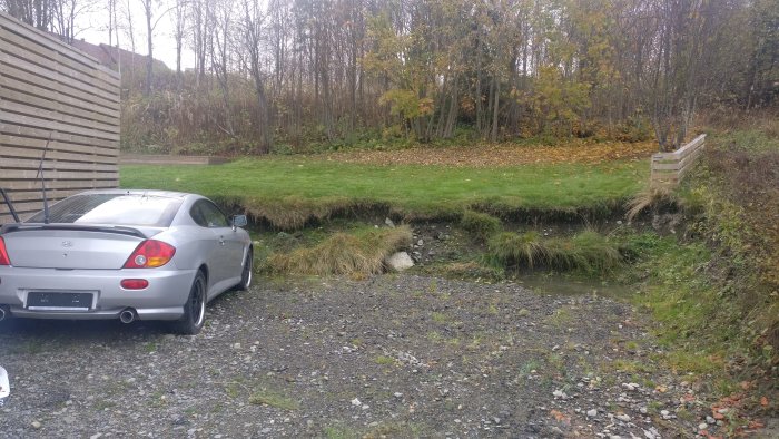 Eroderad kant av gräsmatta vid parkering med mindre grus och grå bil.