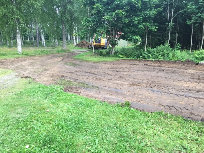 Grävmaskin som förbereder mark för en byggplatta omgiven av grönskande träd och gräs.