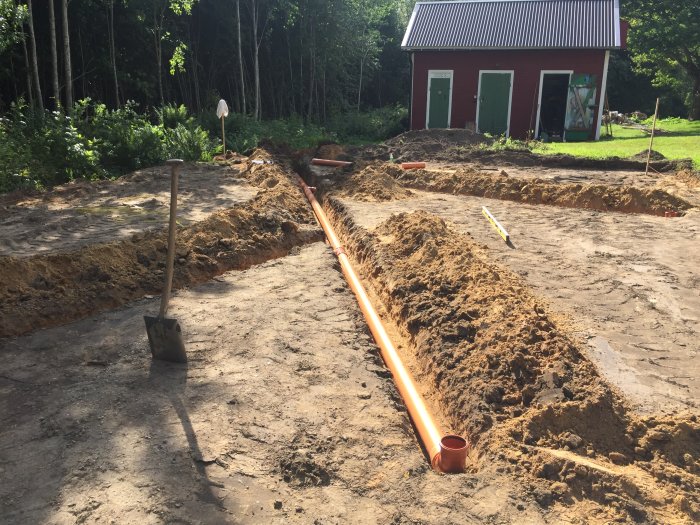 Grundarbete för garage med synliga avloppsrör och spade i framgrunden, röd byggnad i bakgrunden.