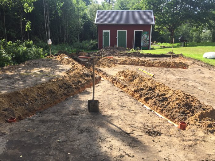 Utgrävning för grund med nedgrävda avloppsrör och spade i förgrunden, med röd ladugårdsbyggnad i bakgrunden.