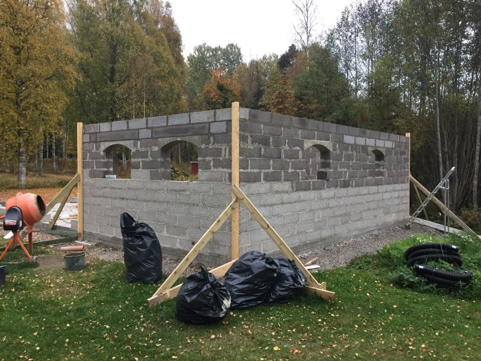 Delvis uppförd grå mur av betongblock med valvformade öppningar för fönster, byggarbetsplats med höstträd i bakgrunden.