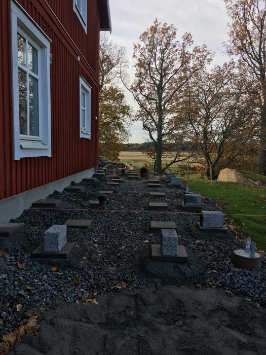 Grundarbete för altan med makadam, riktsnören och gjutna plintar bredvid röd husvägg.