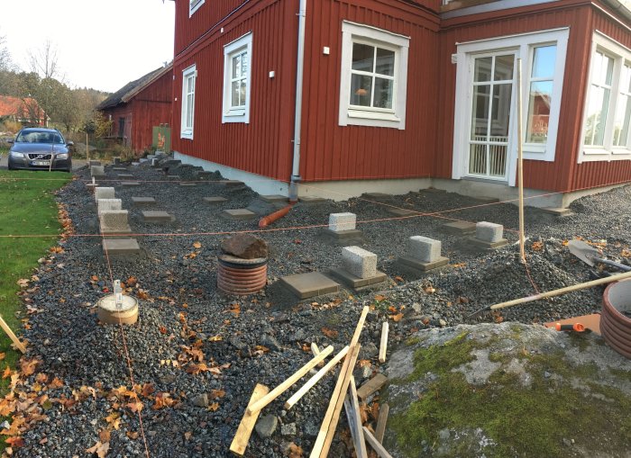 Byggprojekt med makadamgrund, riktsnören, betongplintar och utplacerade stenplattor vid röd trähusfasad.