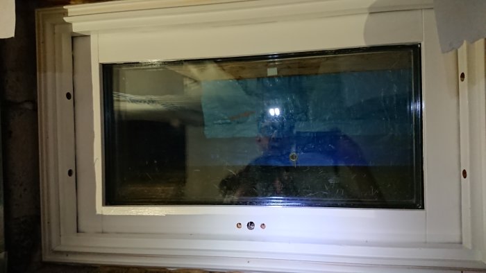 Reflektion av en person med pannlampa som arbetar sent på ett fönsterprojekt, synlig genom ett fönster.