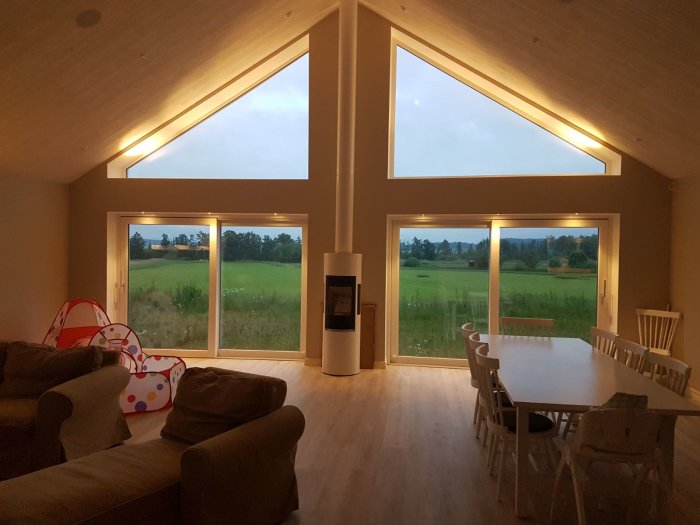 Nytt vardagsrum med stora fönster som vetter mot ett grönt landskap vid skymningen, möbler synliga.
