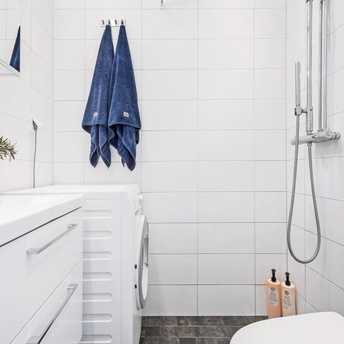 Renoverat badrum med vita kakelväggar, framträdande tvättmaskin och dusch, samt blå handdukar och hygienprodukter.