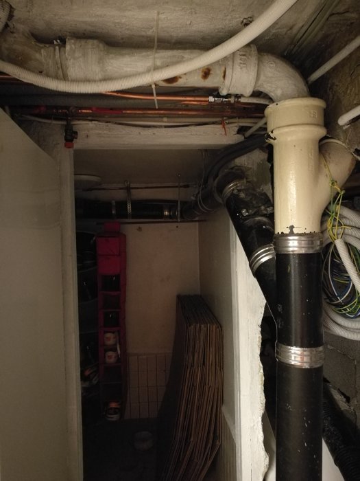 Källarrum med synliga rörinstallationer i taket, inklusive svarta avloppsrör från 2013 och en äldre gul avloppsluftare.