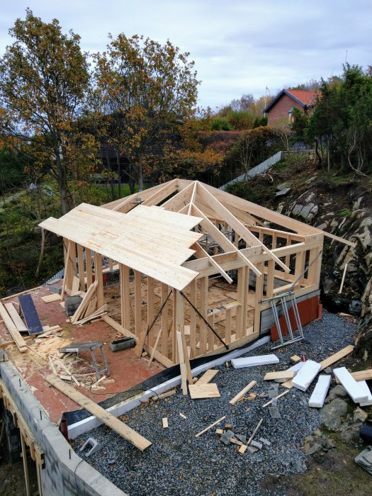 Delvis byggd trästomme till ett hus på en grusgrund med byggmaterial och verktyg utspridda runtomkring.