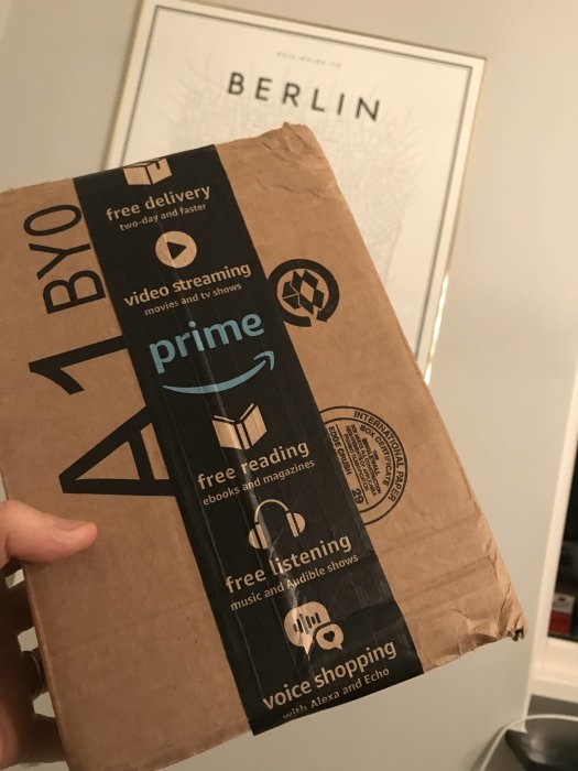 En hand som håller ett brunt Amazon Prime-paket med logotyp och tjänsteerbjudanden.