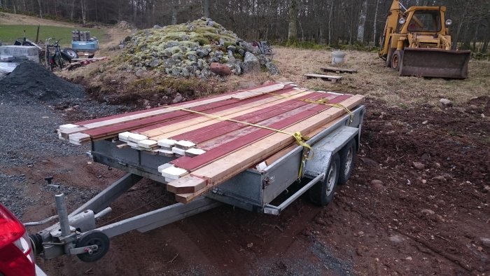 Släpvagn lastad med träplank och isolermaterial för bygge av hönshus, med grävmaskin i bakgrunden.