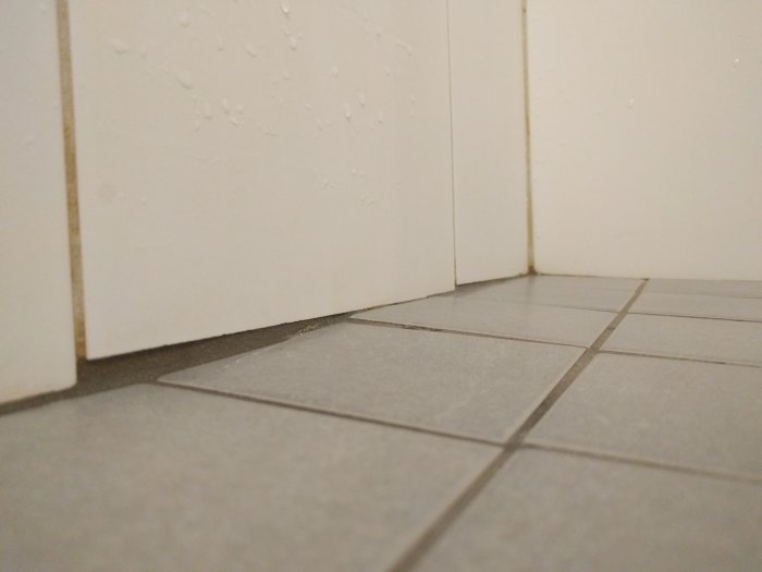 Sprickor i badrumskakel på golvet nära väggen, indikerar tryckskada från underlaget.