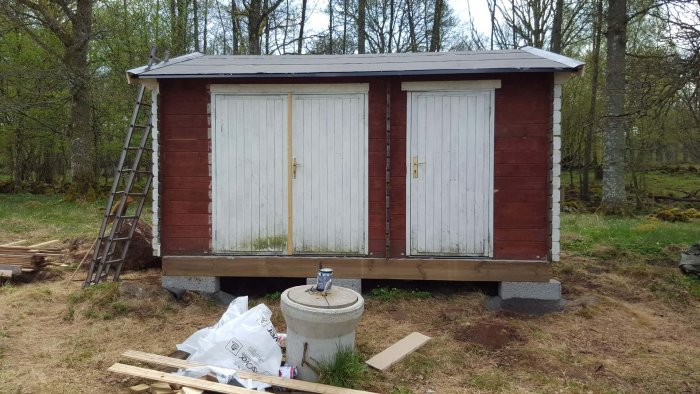 Ett nymålat hönshus med röda väggar och gråa dörrar, uppställt på en äng med skog i bakgrunden.
