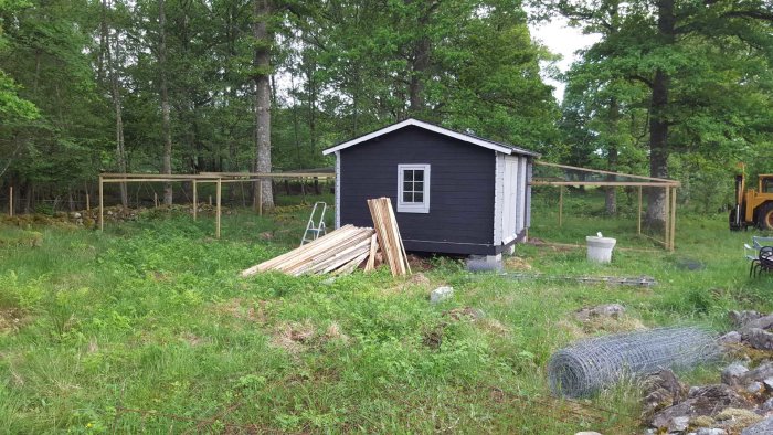 Svartmålat hönshus med gråa dörrar och delar av en hönsgård under konstruktion omgivet av grönskande natur.