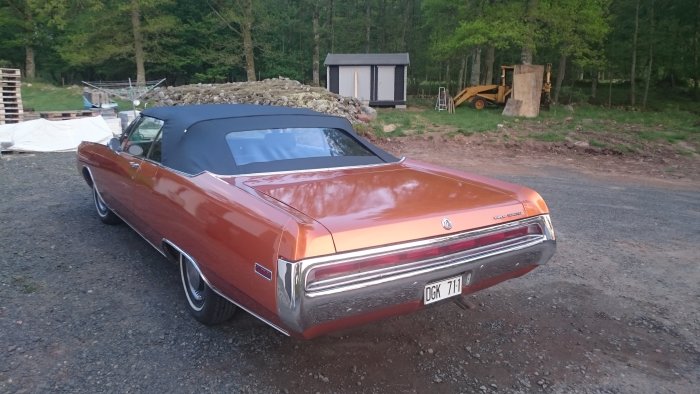Orange Chrysler 300 cabriolet framför ett svart hönshus med gråa dörrar i en lantlig miljö.