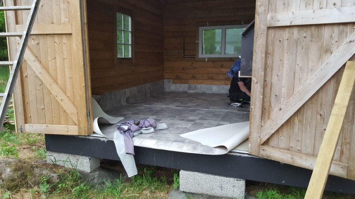 Träkonstruktion av ett väderskyddat hönshus under uppbyggnad med öppna dörrar och inlagd matta.
