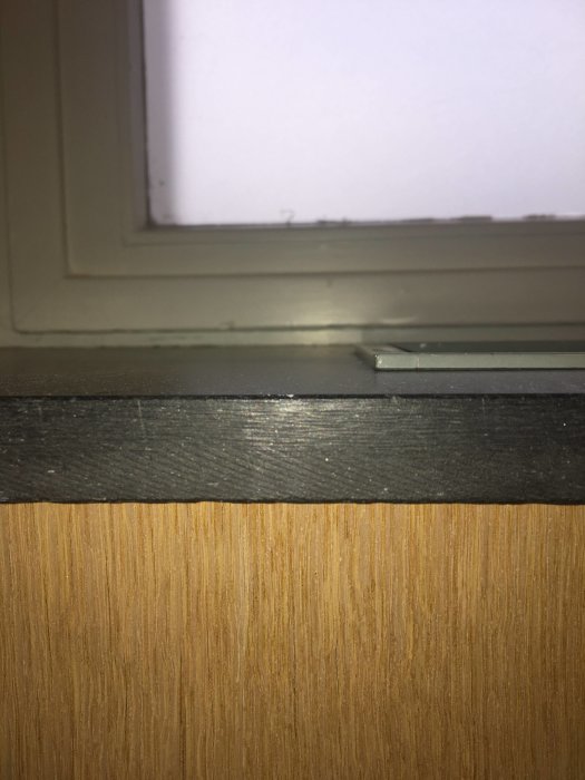 Fönster med svart fönsterbänk ovanför en träyta, misstänkt vara Eternitskiva.