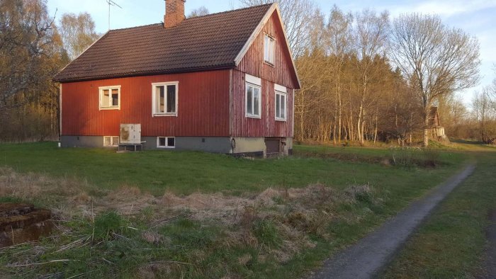 Ett rött trähus med svart tak omgivet av gräsmark och skog i gyllene kvällsljus.