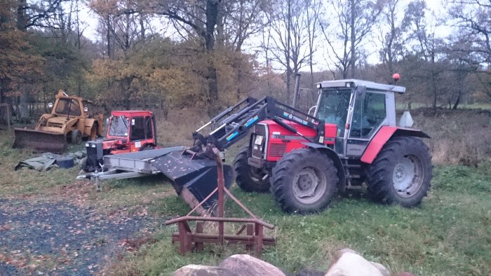 Traktor med frontlastare och släp bredvid en rostig grävmaskin på ödet lantligt område.