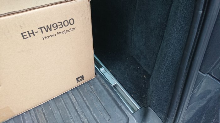 Projektorförpackning i ett bilbagage med synlig kabel och väggfäste, ingen tv installerad.