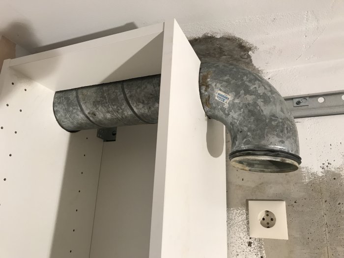 Överskåp anpassat för ventilation med urkapning, bredvid oanvänd eluttag på betongvägg.