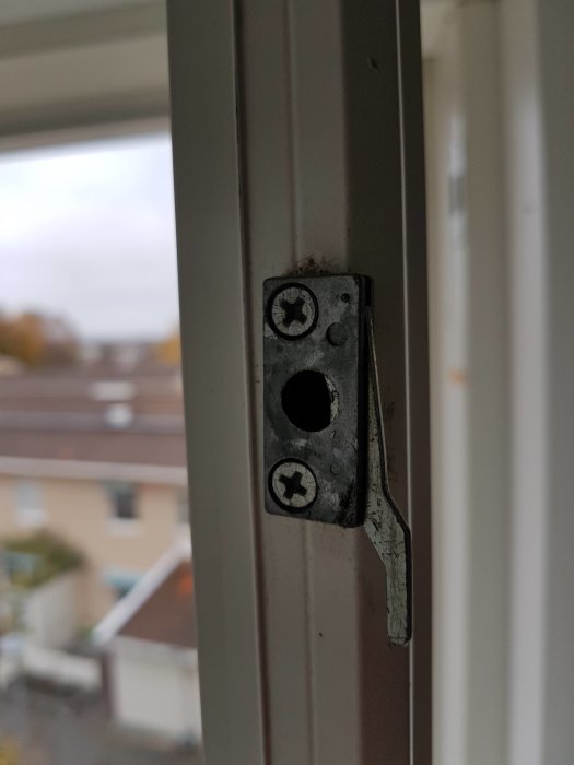 Slitet fönsterlås på en vit fönsterkarm med utsikt mot bostadsområde i bakgrunden.