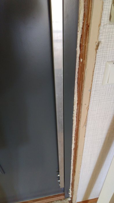 Metallrätskiva mot dörrkarmen visar att karmen böjer inåt nära andra skruven uppifrån.